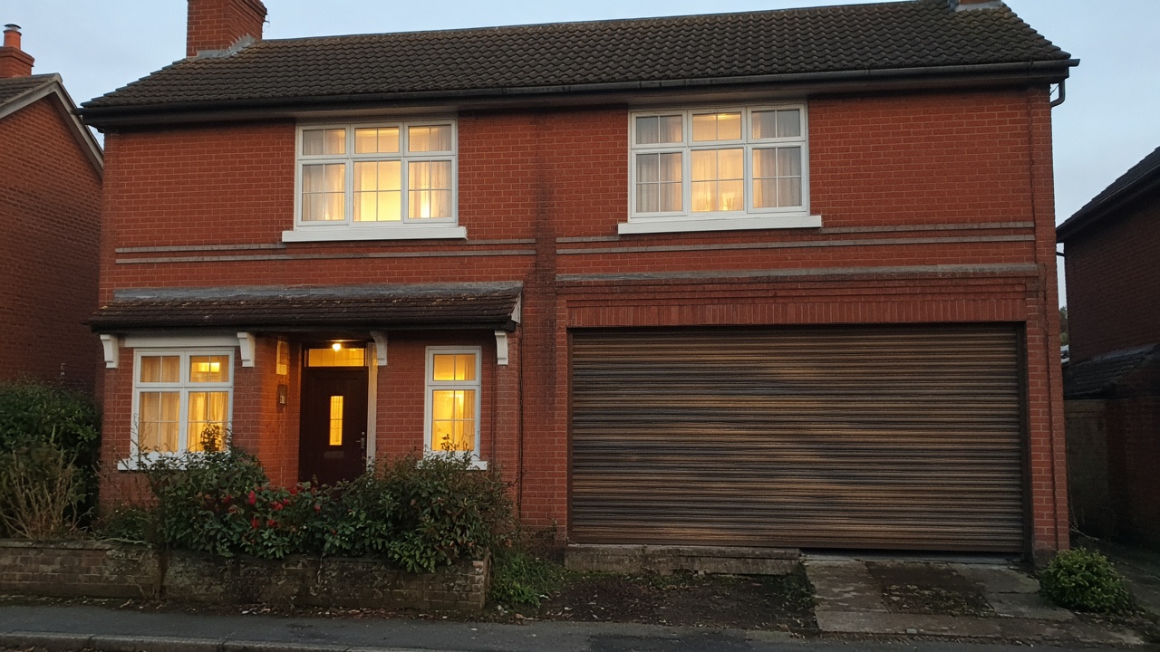 A bronze roller shutter nearly closed on a 1930s red-brick semi, dusk, with warm lights on inside. Wide view of the whole elevation.