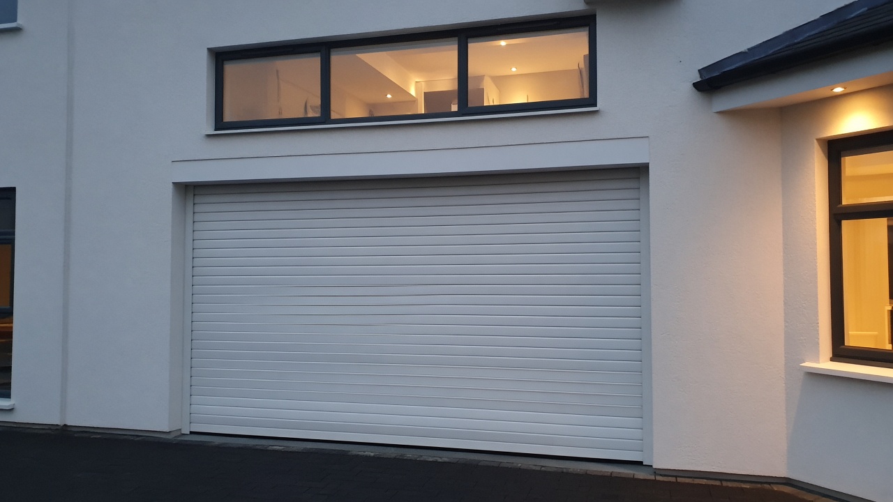 A white roller shutter fully lowered on a white rendered new-build, dusk, with warm lights on inside. Framed on one opening.