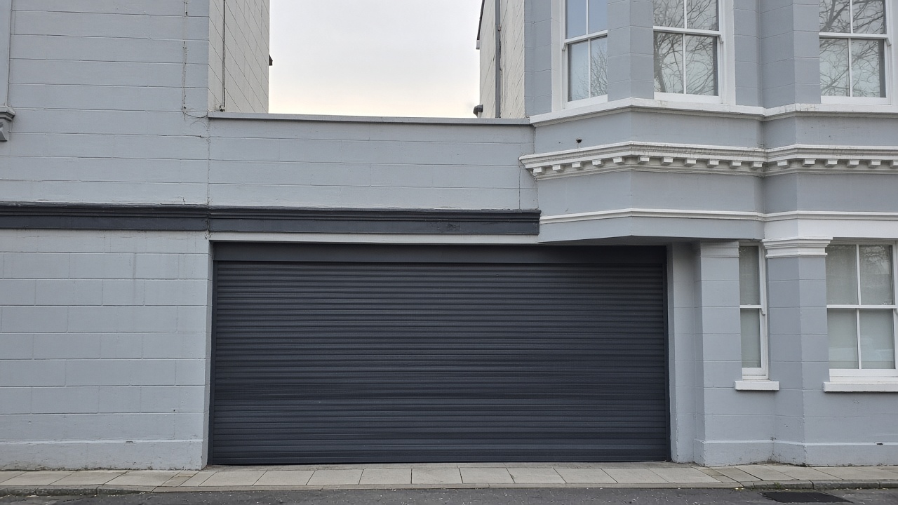 An anthracite roller shutter fully lowered on a painted townhouse with a bay, a flat winter sky. Framed on one opening.